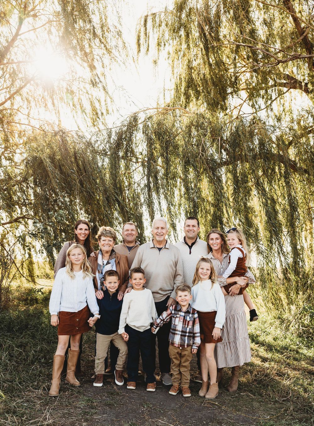 A multi-generational family poses together under a weeping willow tree on a sunny autumn day.