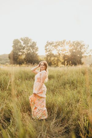 Young woman in floral maxi dress standing in golden sunlit field with tall grass and trees at sunset.