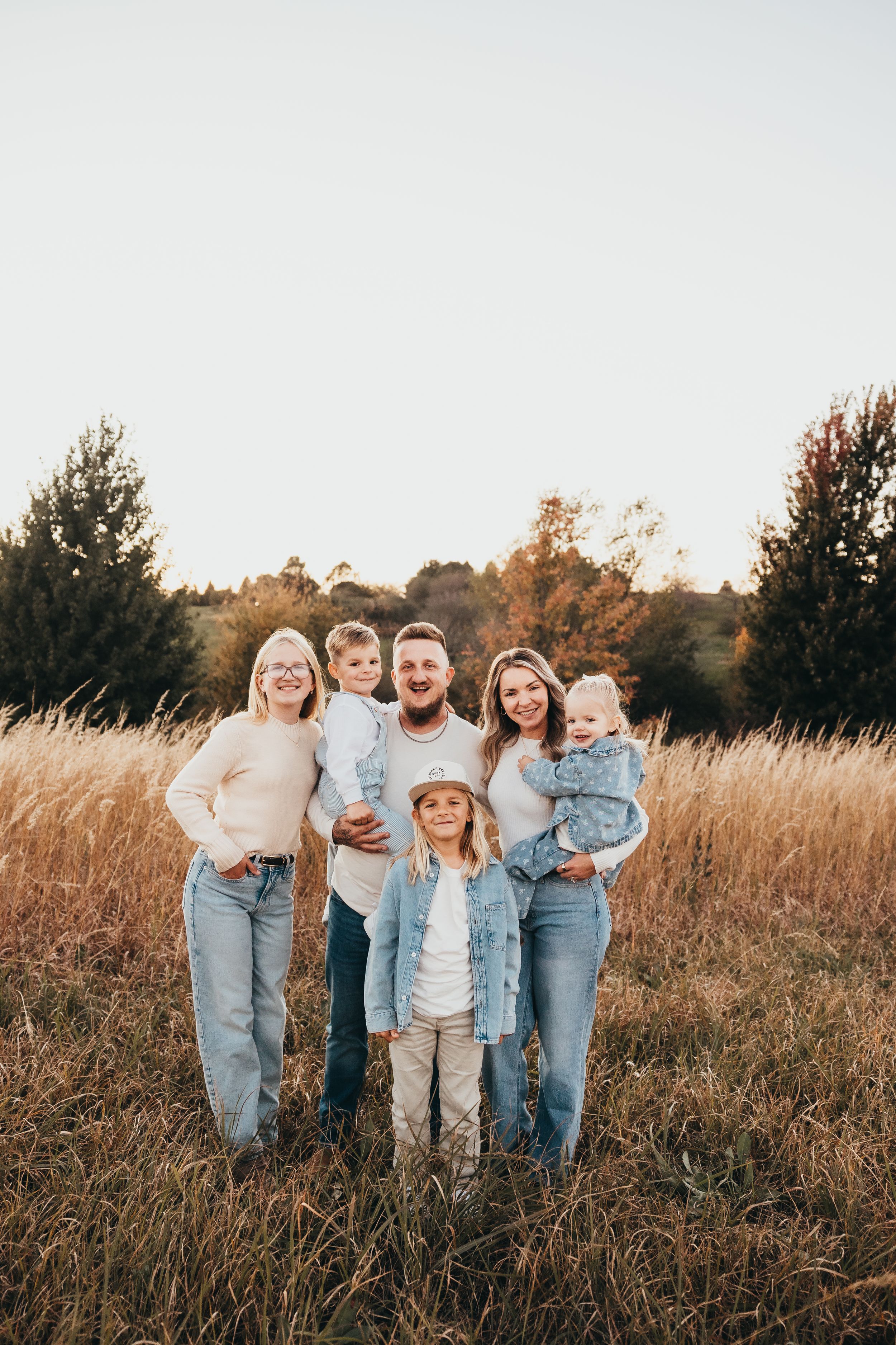 A smiling family of six in cozy neutral outfits poses together in a golden grassy field with trees and a soft sky behind them.
