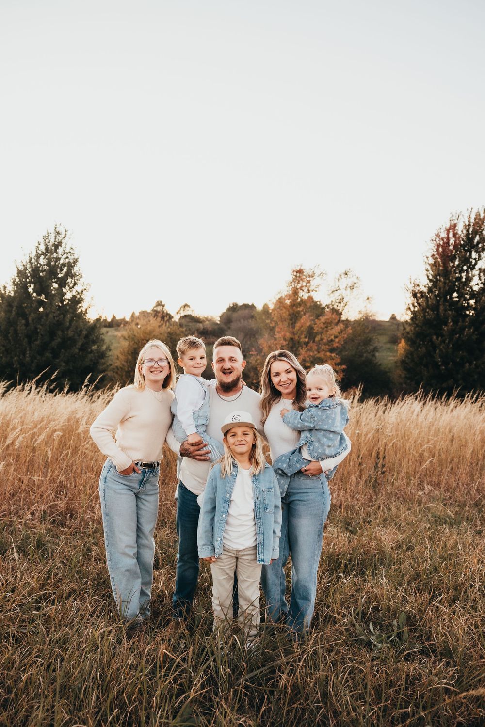 A smiling family of six in cozy neutral outfits poses together in a golden grassy field with trees and a soft sky behind them.