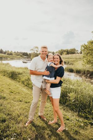 Happy family of three posing outdoors near a river, parents smiling while holding their toddler on a sunny day.