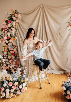 A woman in a floral studio holds a red-haired boy on a ladder, surrounded by colorful flower arrangements.