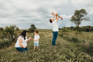 Family enjoying outdoor time in a wildflower meadow, father tossing baby in the air while mother and toddler watch.