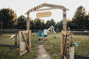 Two children walk hand-in-hand through a rustic wooden farm gate entrance on a sunny day.