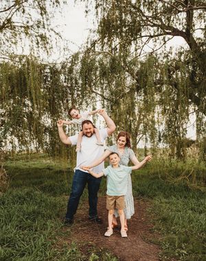 Happy family of four posing under a weeping willow tree, with child on dad's shoulders outdoors.