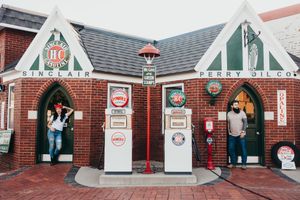 Two people stand beside vintage gas pumps at a restored red brick Sinclair and Perry Oil Co. filling station with gabled roofs.
