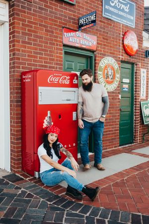 Couple poses by a vintage red Coca-Cola machine outside a brick garage with retro Ford, Sinclair and Prestone signs.