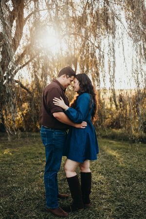Couple embracing forehead to forehead under a weeping willow tree at golden hour during an outdoor engagement photo session.