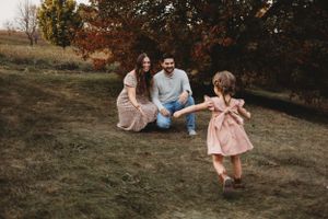 A young child in a pink dress twirls in an autumn field while adults sit and watch on the grass.