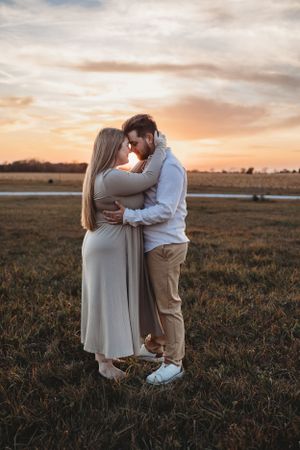 A romantic couple embraces in a field during golden hour sunset with warm autumn colors in the background.