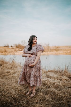 A person in a floral dress stands by a lake during golden hour, cradling their pregnant belly in a serene outdoor setting.