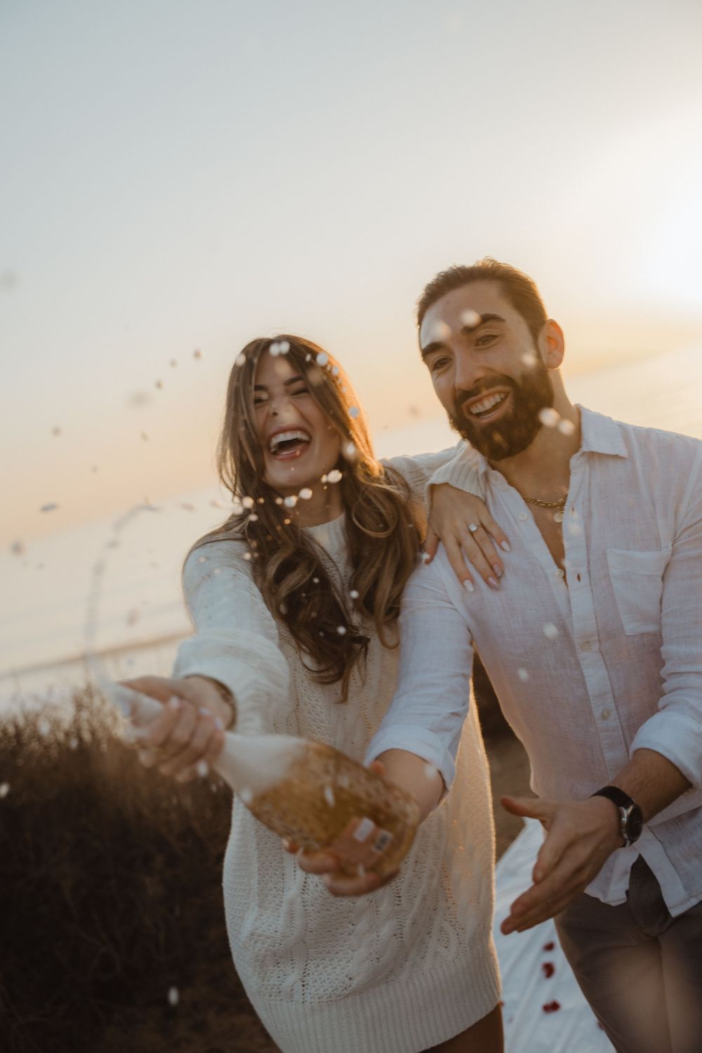 Happy couple laughs joyfully while throwing confetti in warm sunset light.