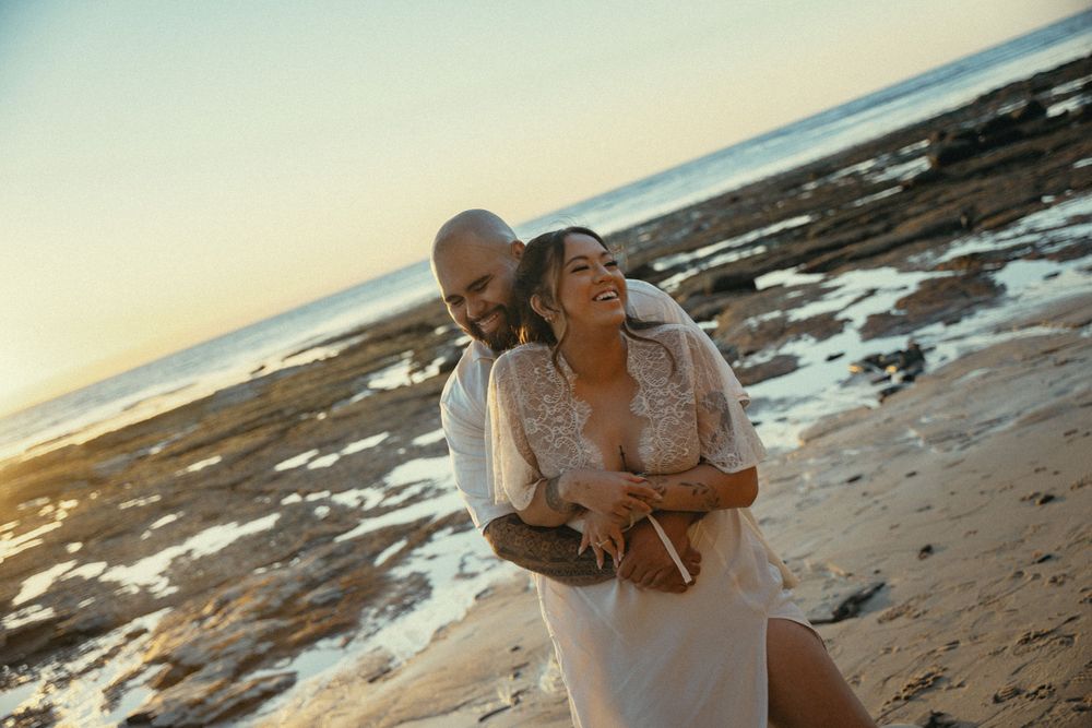 A couple embraces on a beach at sunset while dressed in white outfits.