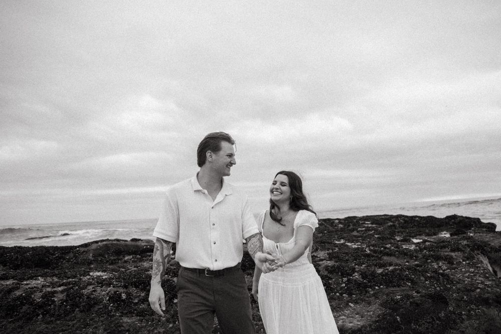 A black and white coastal portrait of a couple on rocky shoreline with cloudy skies.