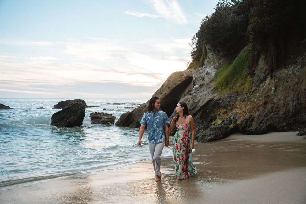 A couple strolling along a scenic beach with cliffs and ocean waves at sunset.