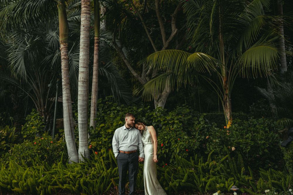 A couple in wedding attire stands among tropical palm trees and lush greenery at dusk.