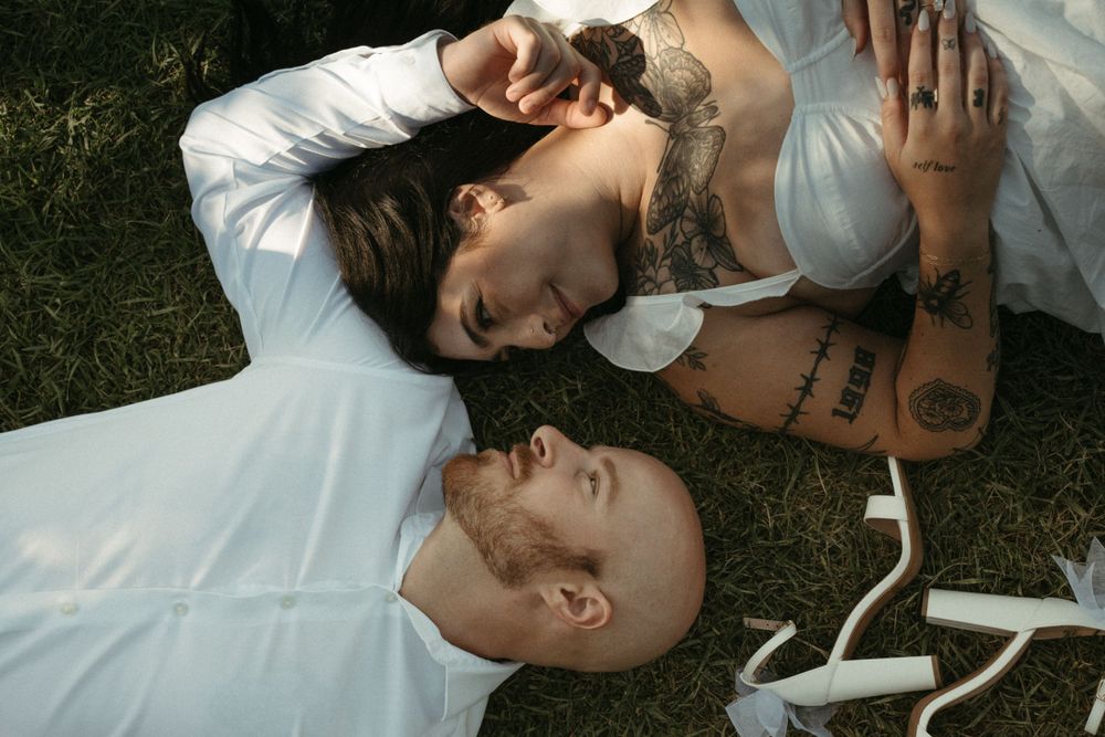 A couple lies on grass together in white clothing during a photo session.