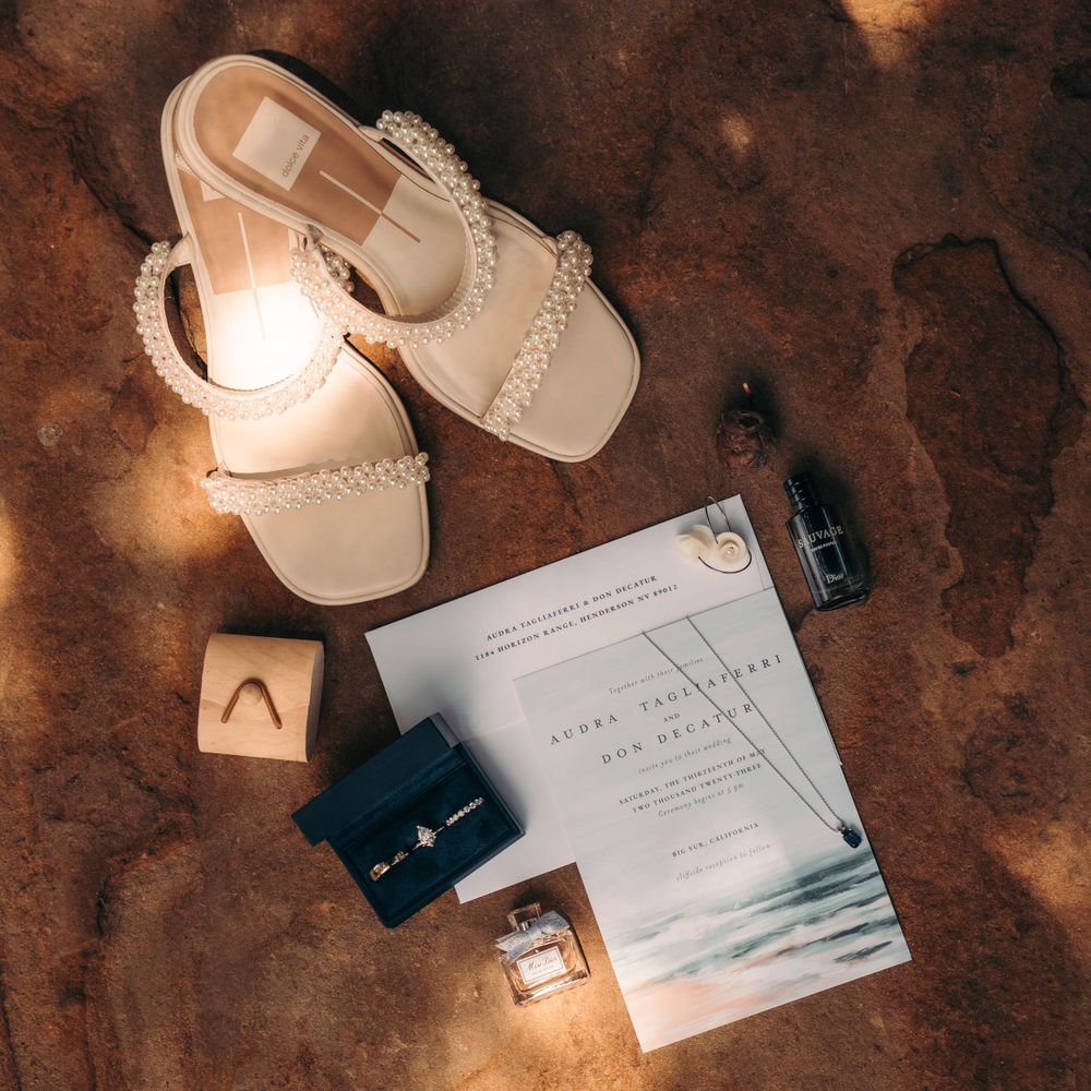 Flatlay of cream sandals, small bottles, and a book on rustic brown surface with warm lighting effect.