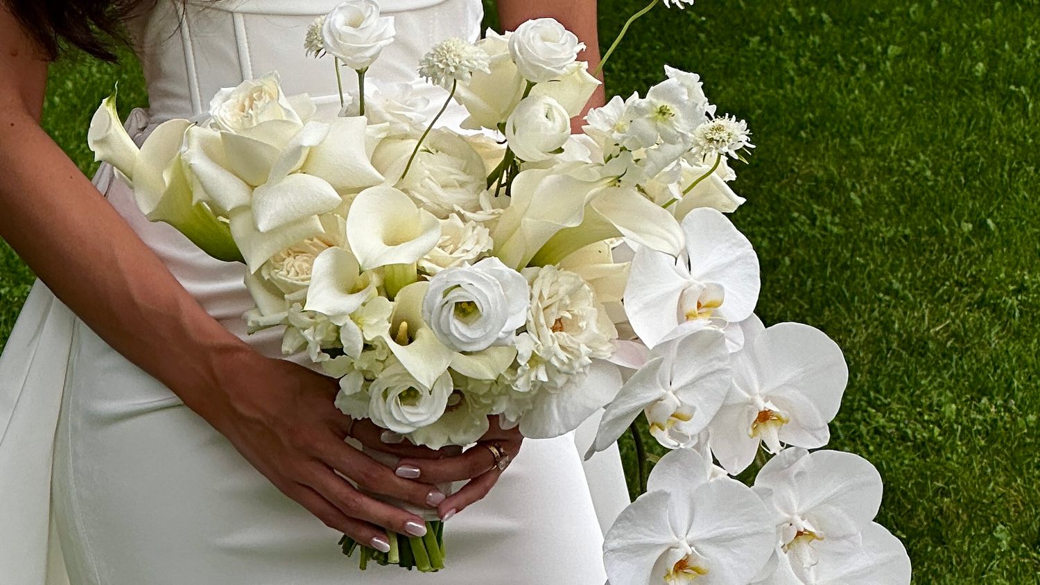 Elegant white wedding bouquet featuring calla lilies, roses, and orchids against green background.