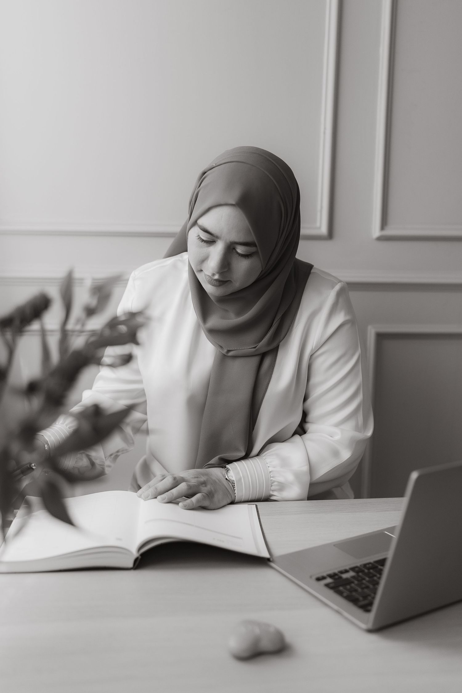 Professional in white hijab and blazer works diligently at desk with laptop and books in monochrome office setting.