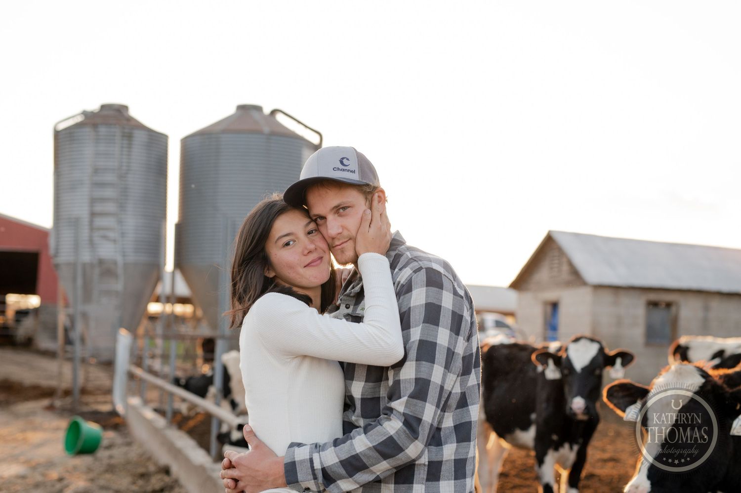 A couple embraces on a dairy farm with silos and cattle in the background during sunset.