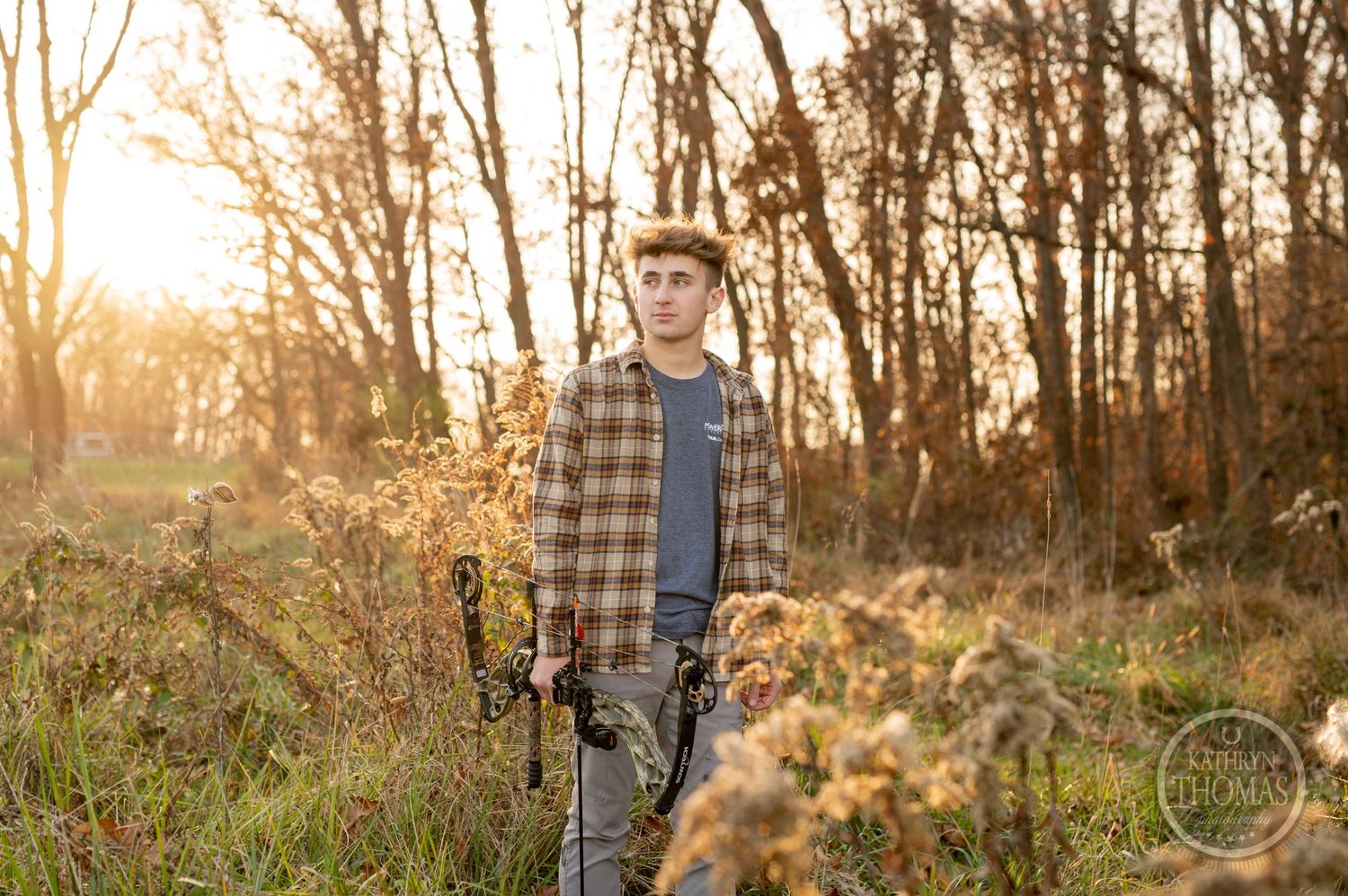Hunter with compound bow stands in golden autumn field at dusk with trees in background.