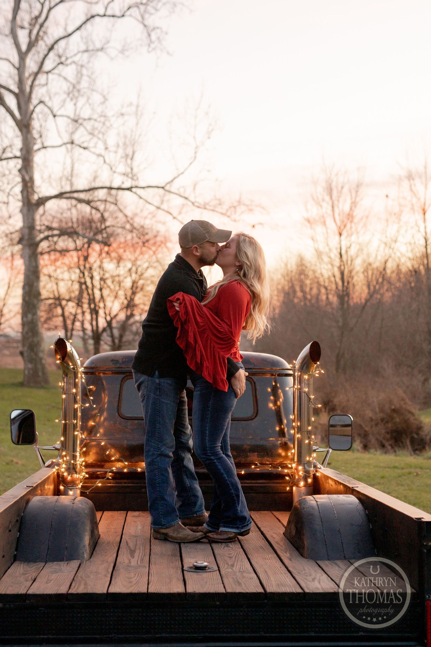 Romantic couple sharing a kiss on a pickup truck bed decorated with string lights at sunset.