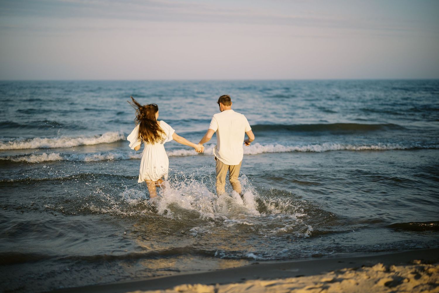 Two people running into ocean waves at sunset wearing white summery outfits and splashing in the water.