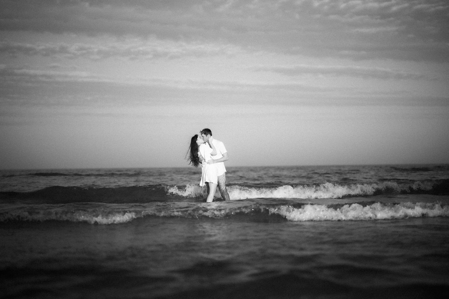 A couple embraces while standing in the ocean waves at sunset, captured in black and white photography.