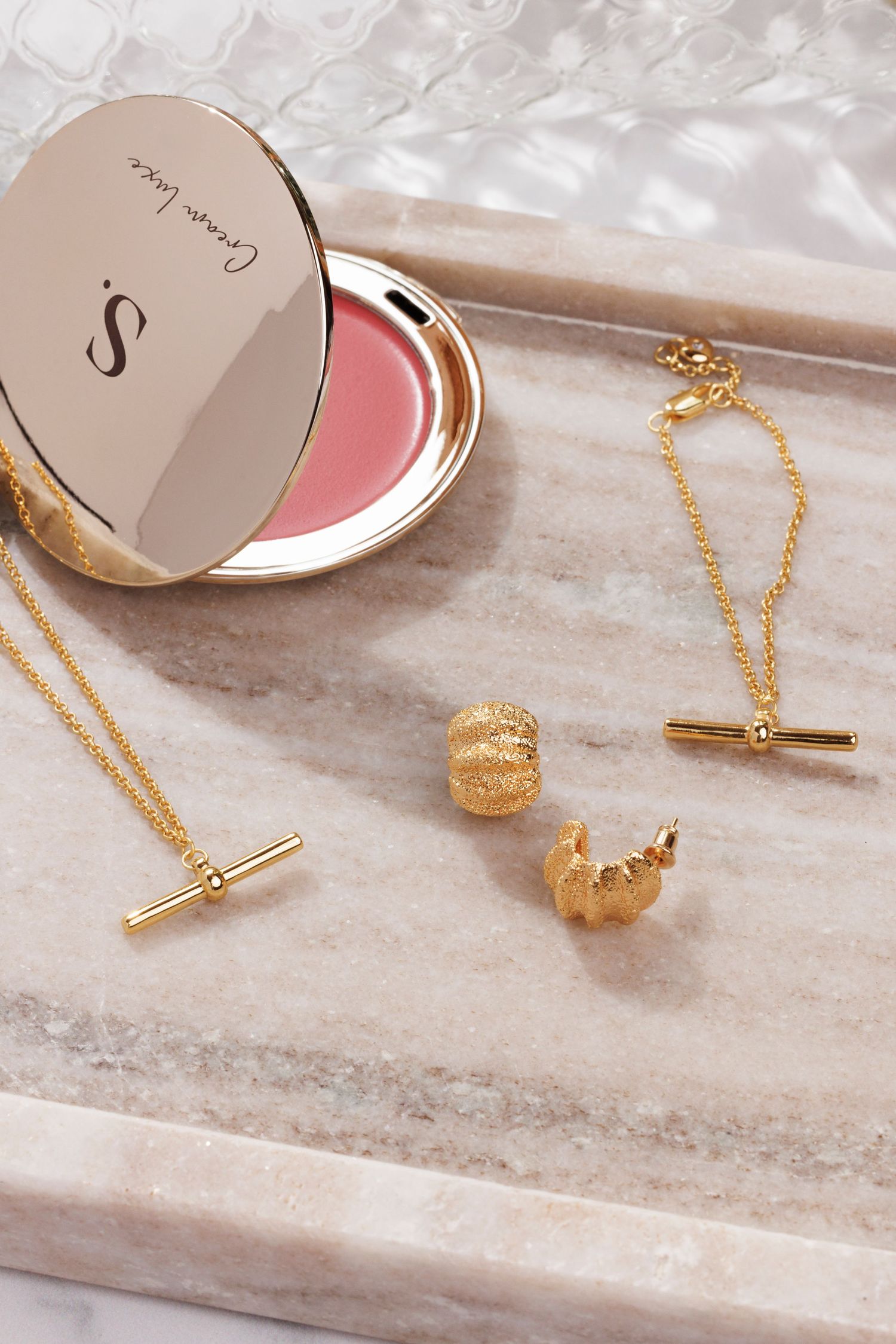 Pink compact blush with gold jewellery and accessories arranged on marble surface. There is glass shadows in the background.