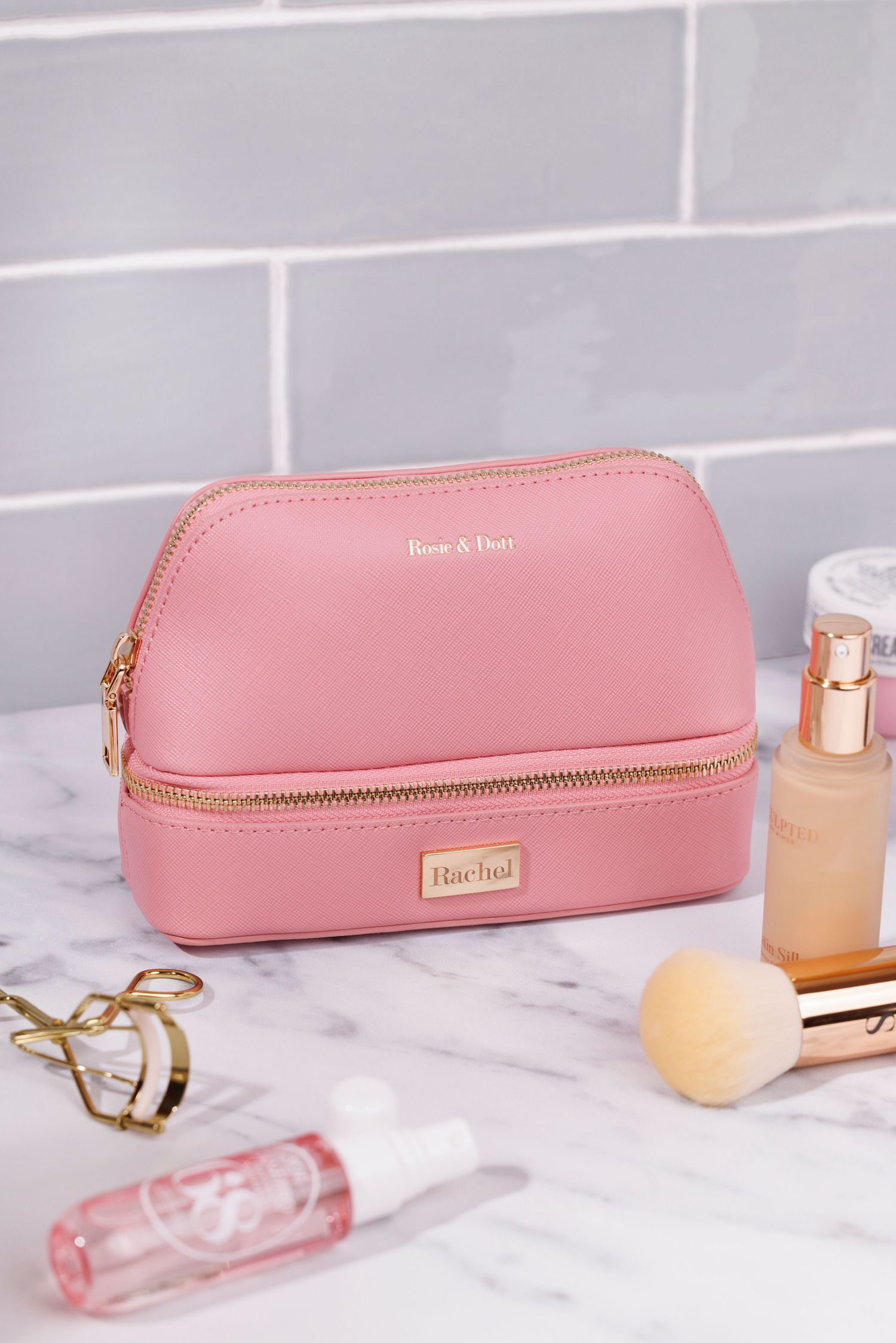 Pink makeup bag with beauty products and accessories on marble counter with a tiled bathroom background.