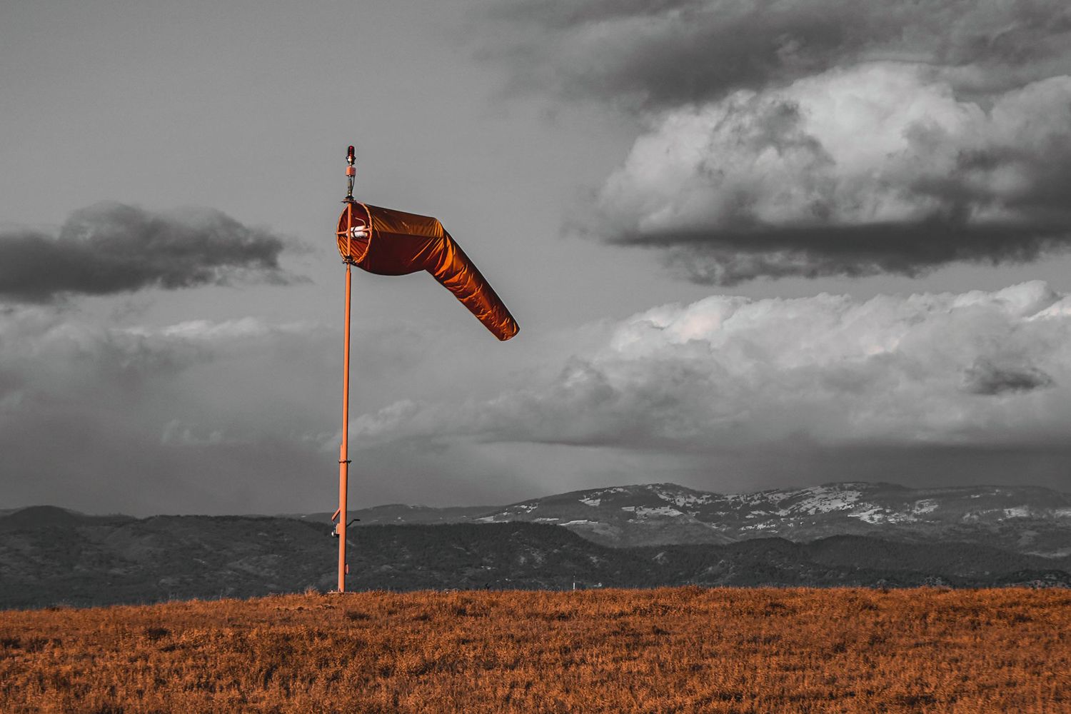 Wind sock monitoring wind conditions against dramatic cloudy sky and mountains in selective color photograph.