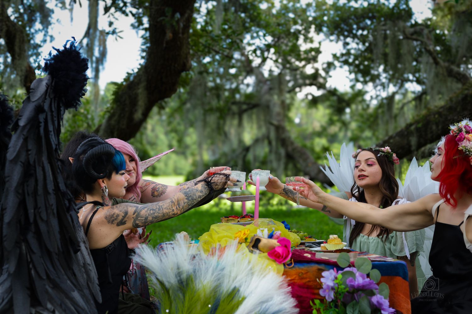 Fantasy-costumed women reaching across a colorful decorated outdoor table under oak trees.