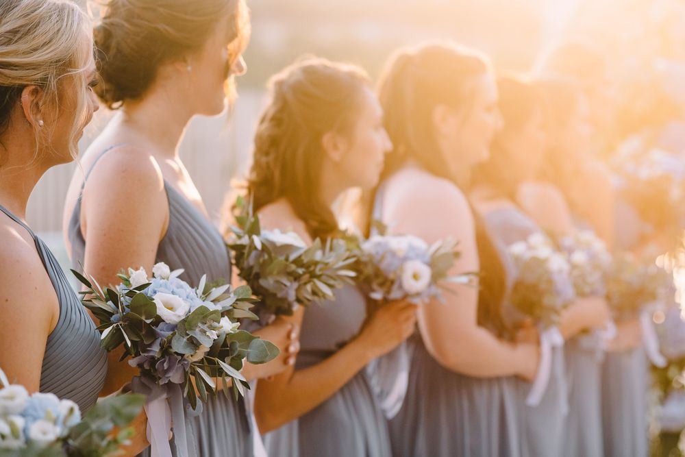 Bridesmaids in gray dresses holding white floral bouquets lined up in golden sunset light.