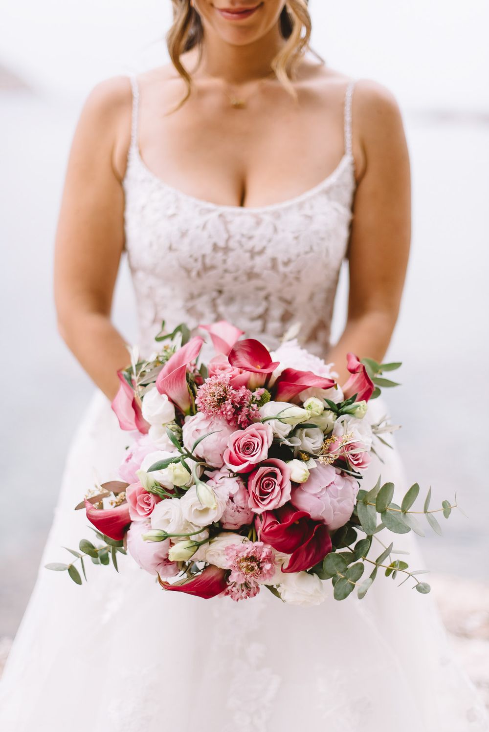 Bride in lace wedding gown holding a lush bouquet of pink roses, peonies, and eucalyptus near water.