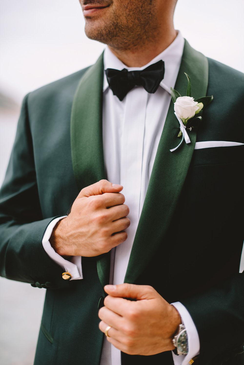 Groom in green tuxedo with black bow tie and white boutonniere adjusting his jacket on wedding day.