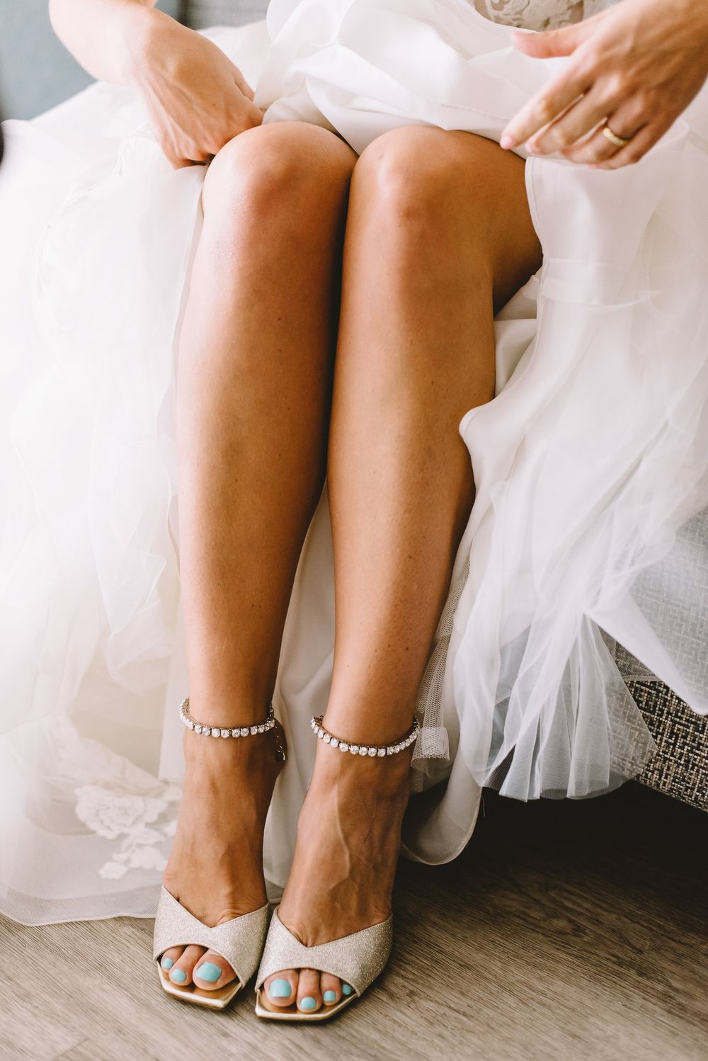 Bride wearing elegant white heeled sandals with pearl ankle straps and blue pedicure, sitting in wedding dress.