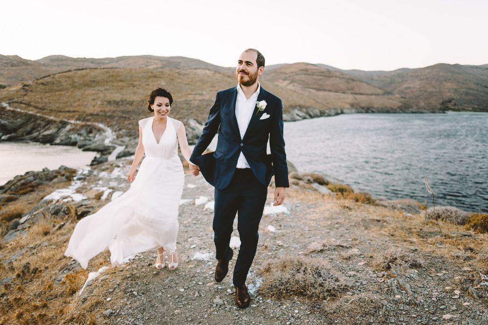 A bride and groom hold hands while walking along a scenic coastal hillside on their wedding day in Greece.