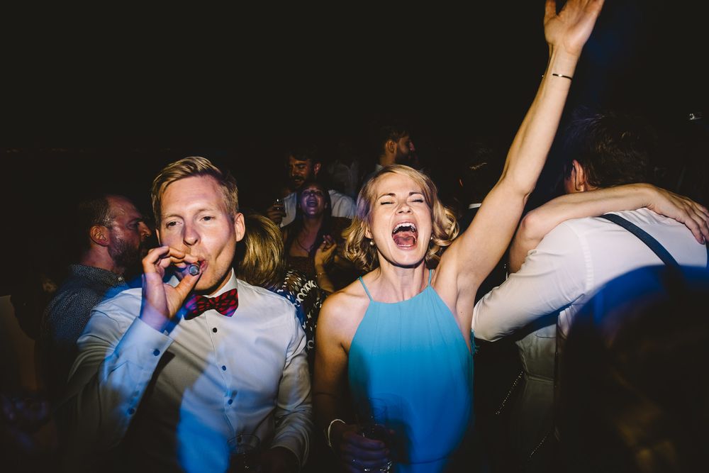 Joyful wedding guests dancing and celebrating at a lively reception party, woman with arm raised laughing.
