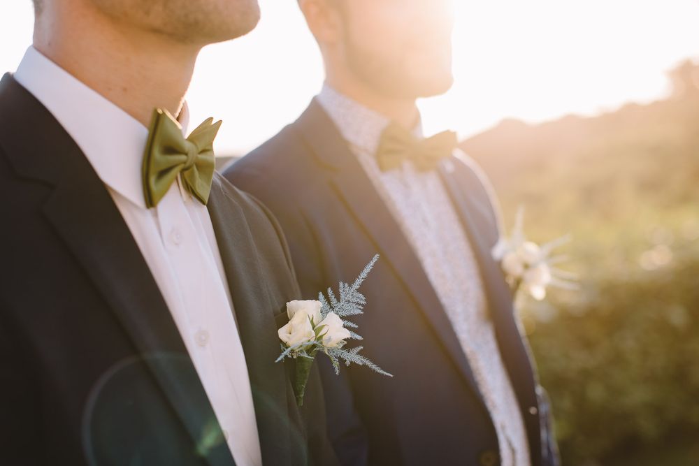 Two grooms in navy suits with olive green bow ties and white boutonnieres stand together in golden sunset light.