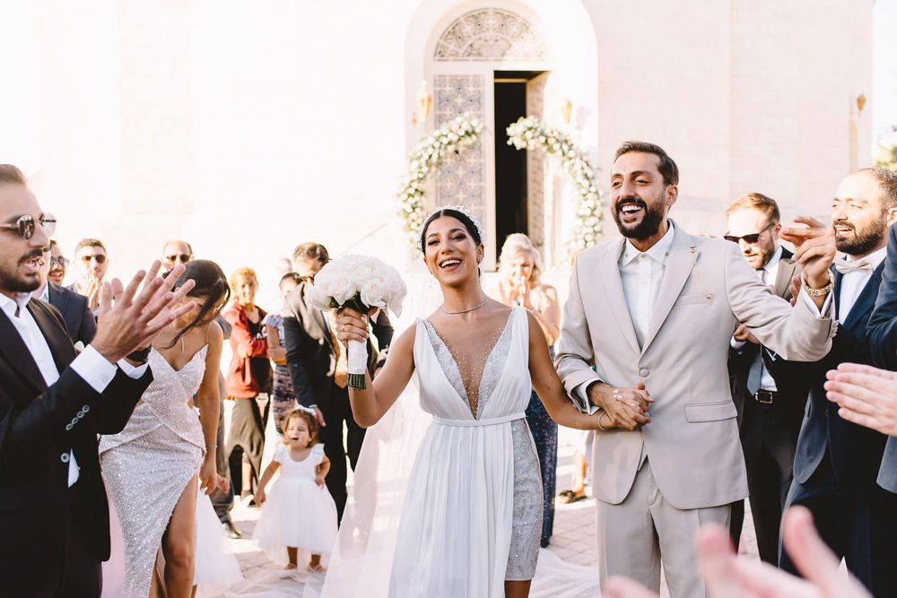 Joyful newlyweds exit church ceremony as guests celebrate with applause, bride in white gown with floral crown.