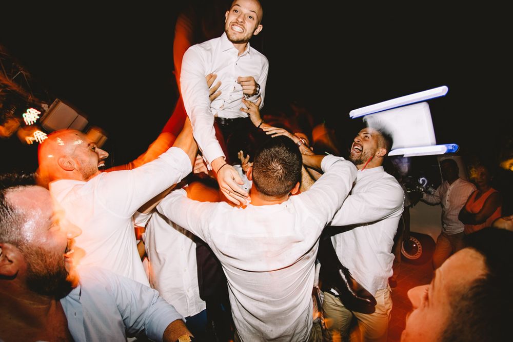 Groom lifted by groomsmen in white shirts during lively outdoor wedding reception celebration at night.