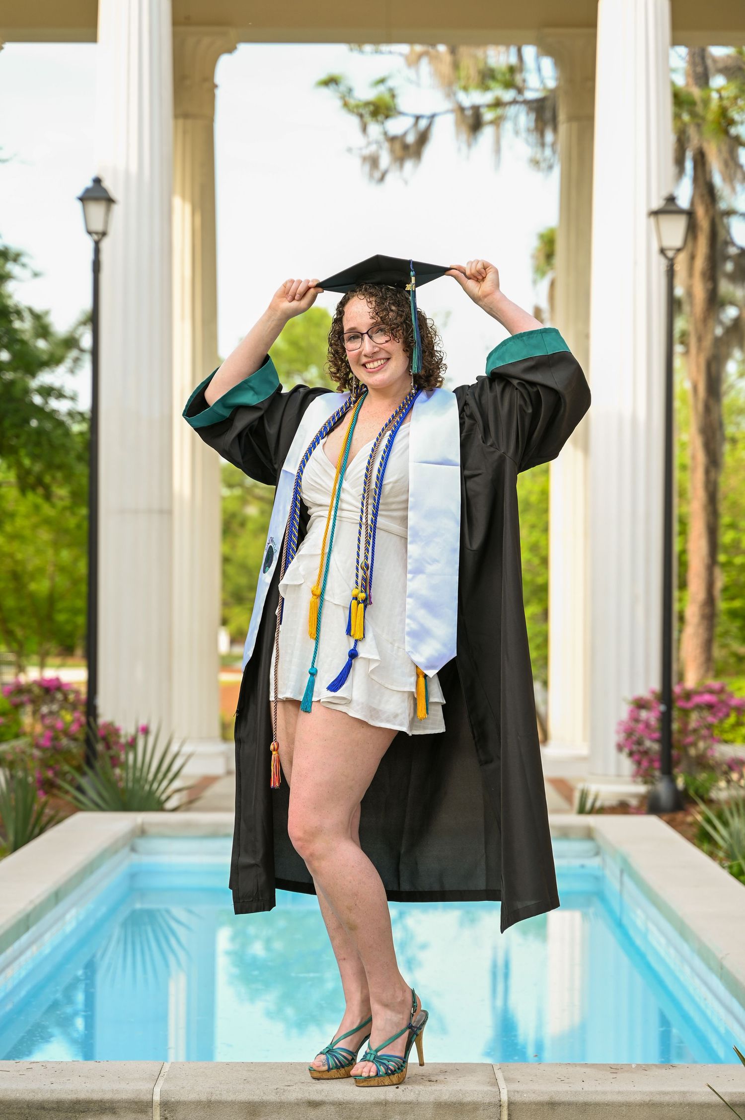 Graduate in cap and gown poses confidently by a swimming pool with white columns.