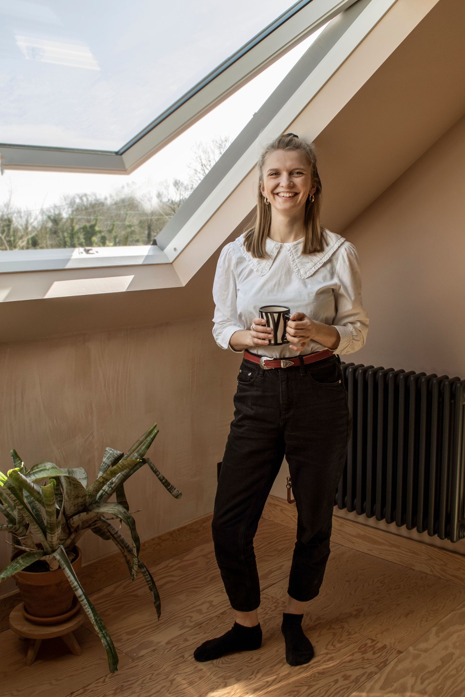 Person in white sweater and dark pants standing by skylight window with indoor plants.