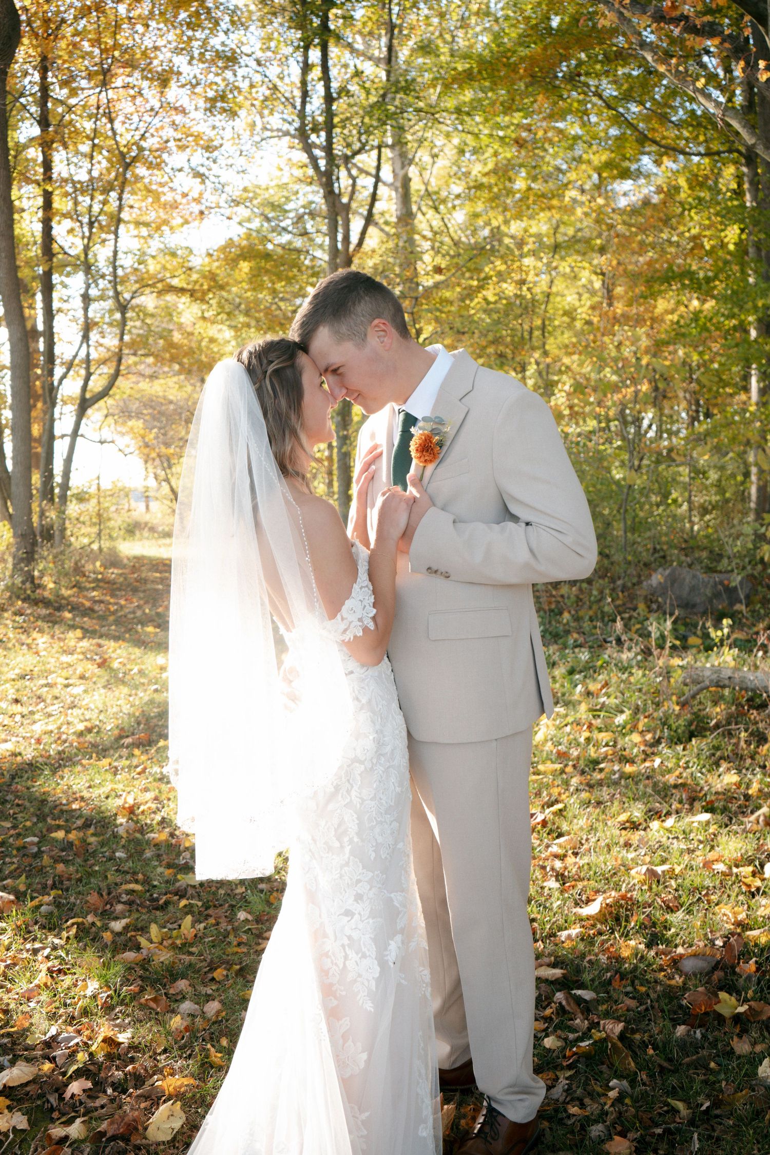 Bride and groom touching foreheads tenderly in golden autumn forest wearing beige suit and lace gown.