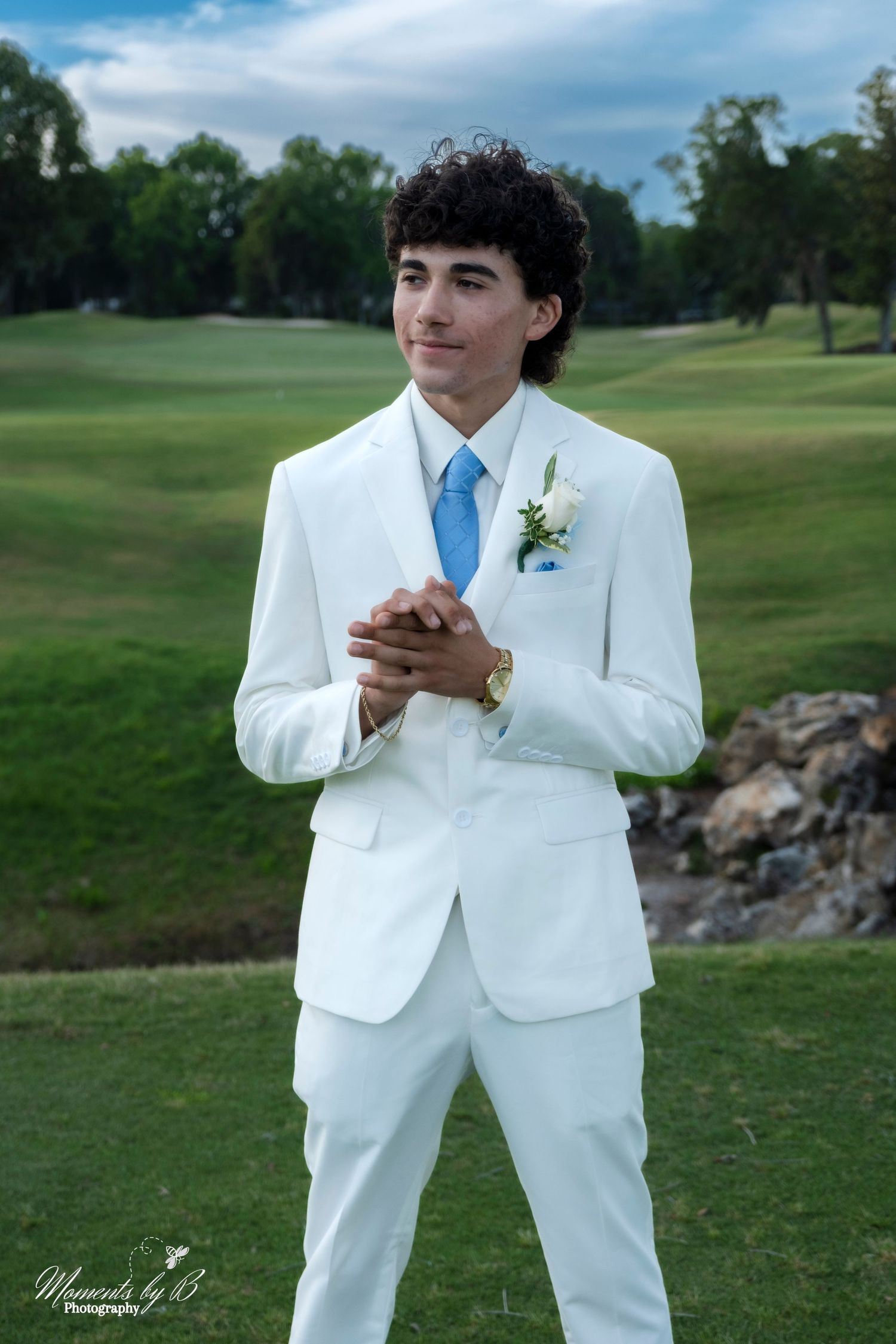 A formal portrait in a white suit with blue tie against a scenic golf course background.
