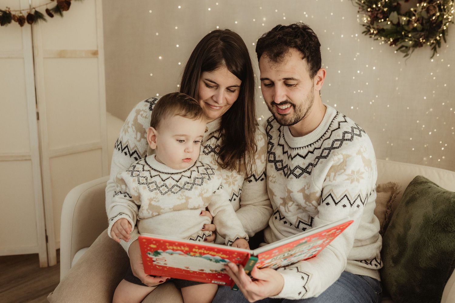 Family in matching winter sweaters reads together on couch with Christmas decorations in background.
