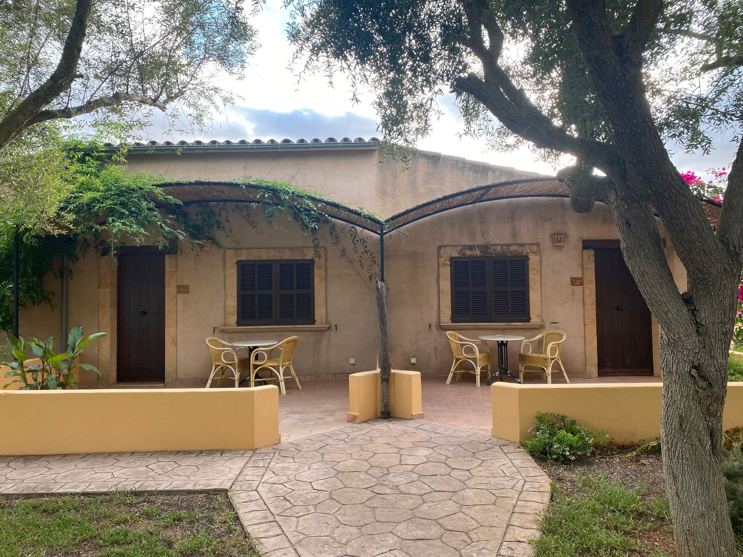 Mediterranean-style bungalow with covered porch and garden paths under shady trees.