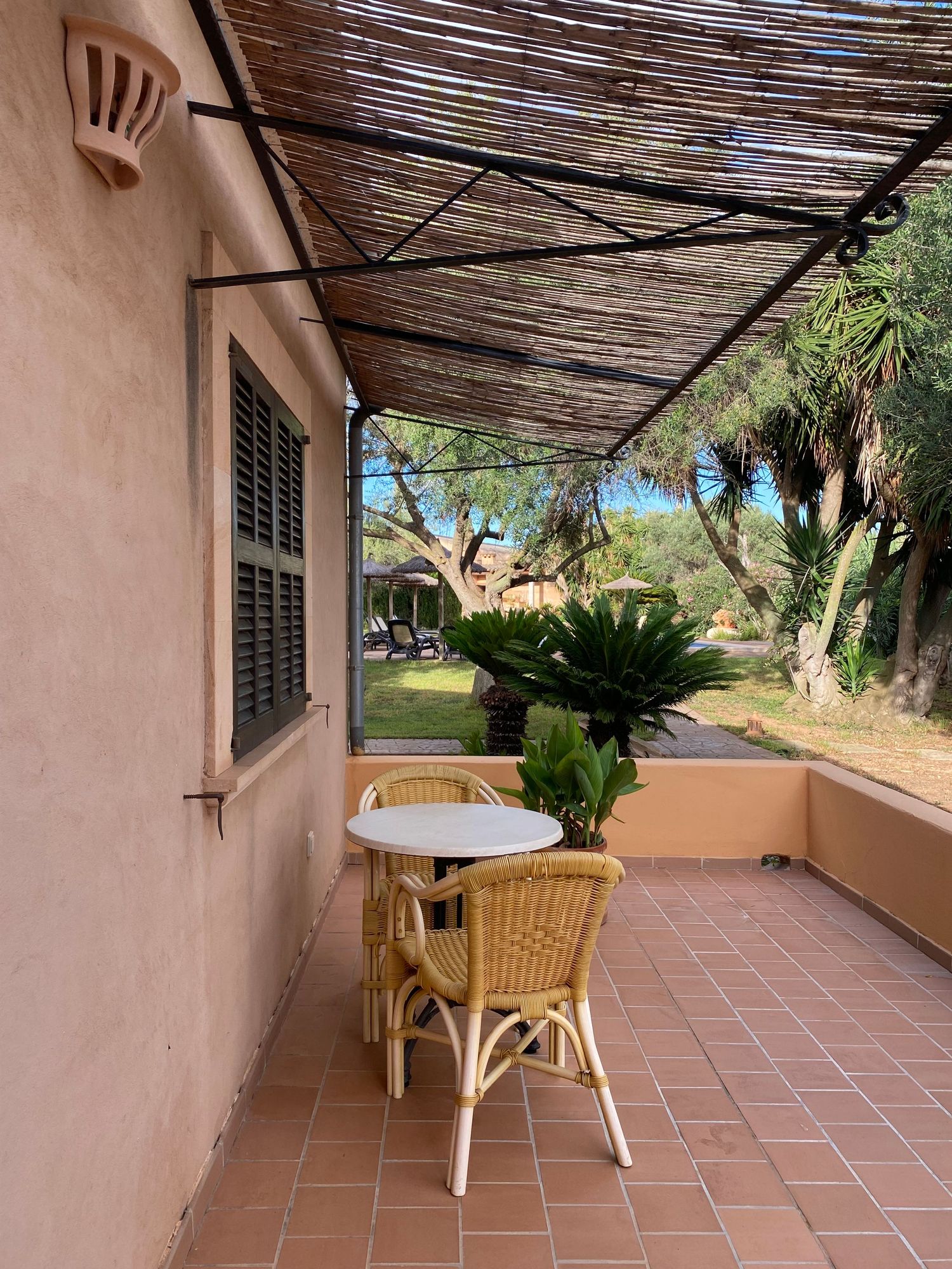 Cozy terrace with wicker chair and table under a rustic bamboo shade cover overlooking a Mediterranean garden.