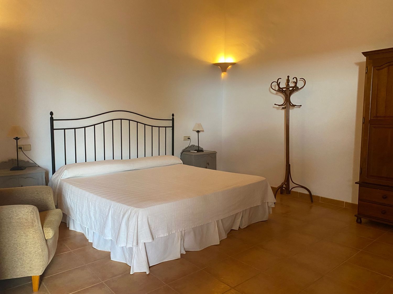 Simple bedroom with white bed linens, iron headboard, and wooden coat stand under warm lighting.